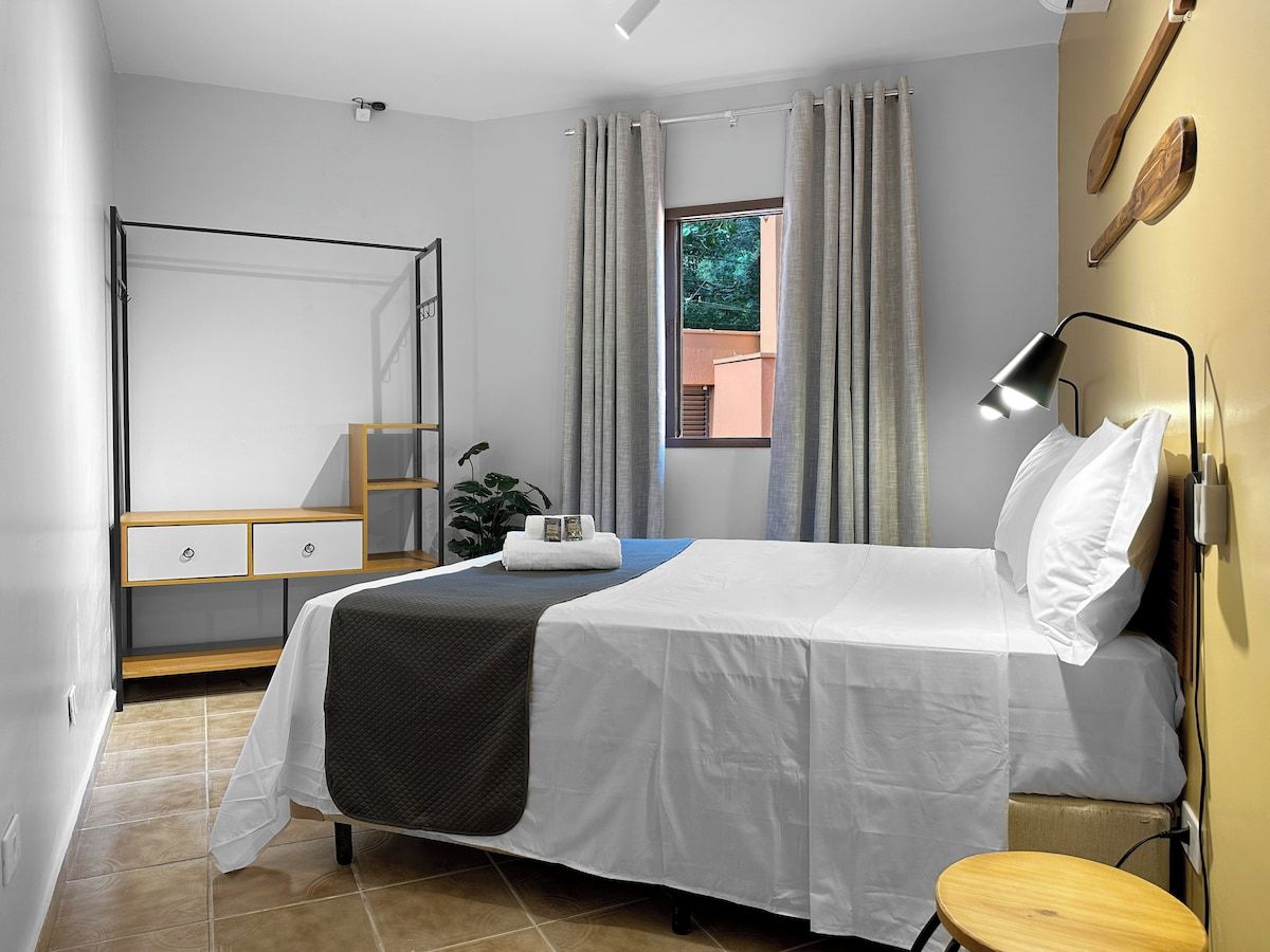 A hotel room with a bed, bedside table, a desk with drawers, a chair, a potted plant, and a window with curtains.