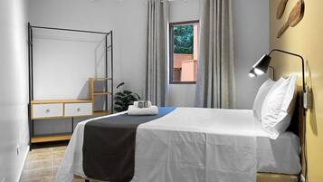 A hotel room with a bed, bedside table, a desk with drawers, a chair, a potted plant, and a window with curtains.