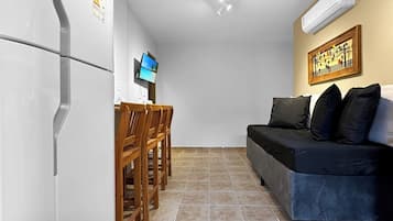 Um espaço de estar compacto com cozinha compacta, área de jantar com cadeiras de madeira, sofá com almofadas e piso ladrilhado.