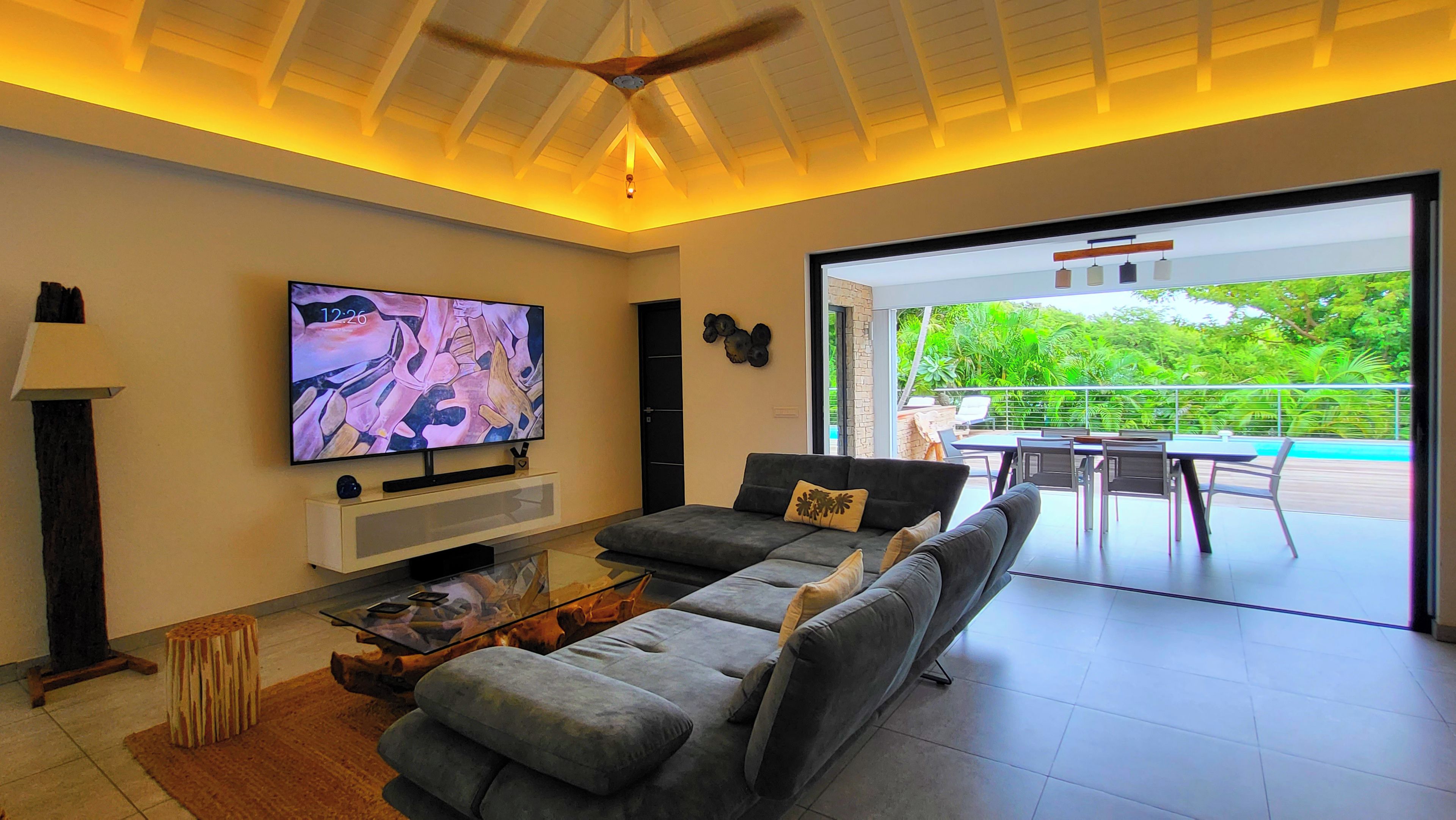A modern living room with a flat-screen TV, a sofa, a coffee table, and a dining area with a view of greenery.