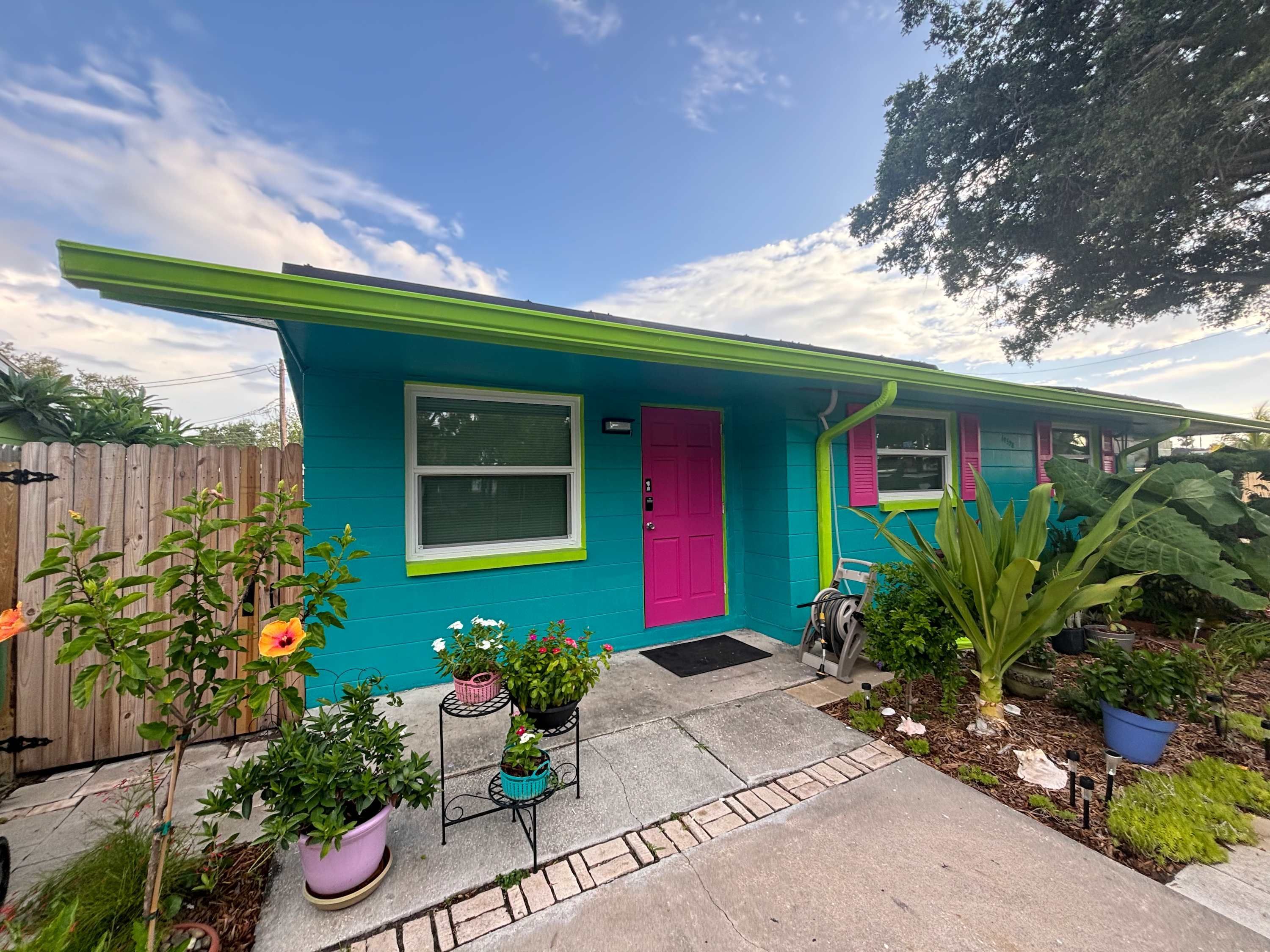Une maison à la façade turquoise, avec une porte rose et une fenêtre dotée d’un cadre blanc. Des plantes en pot sont disposées devant la maison.