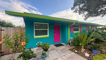 Une maison à la façade turquoise, avec une porte rose et une fenêtre dotée d’un cadre blanc. Des plantes en pot sont disposées devant la maison.
