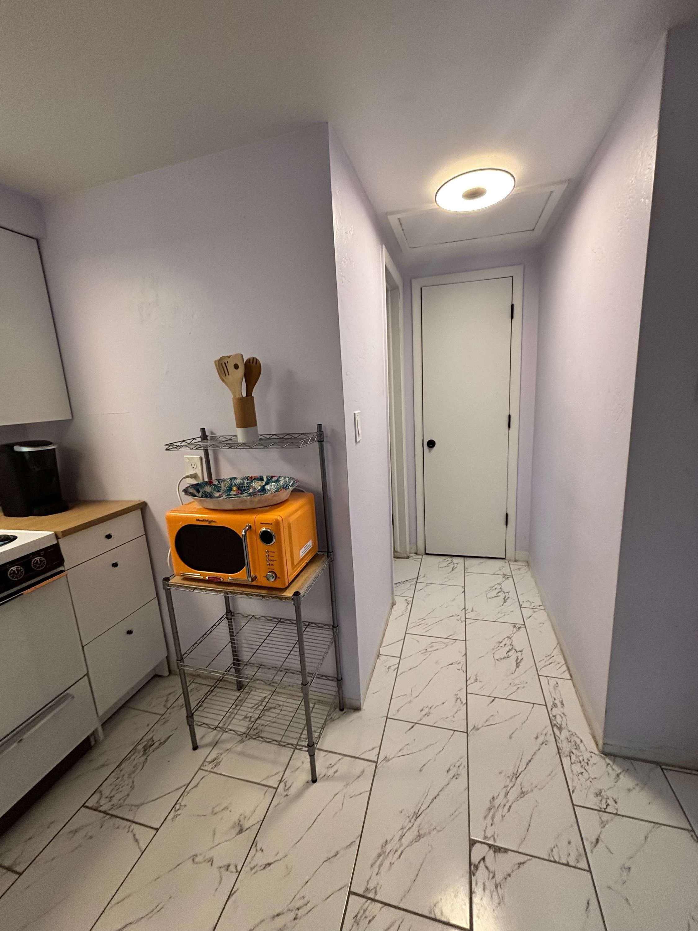 A narrow hallway with a microwave on a metal stand, a stove, and a white door at the end.