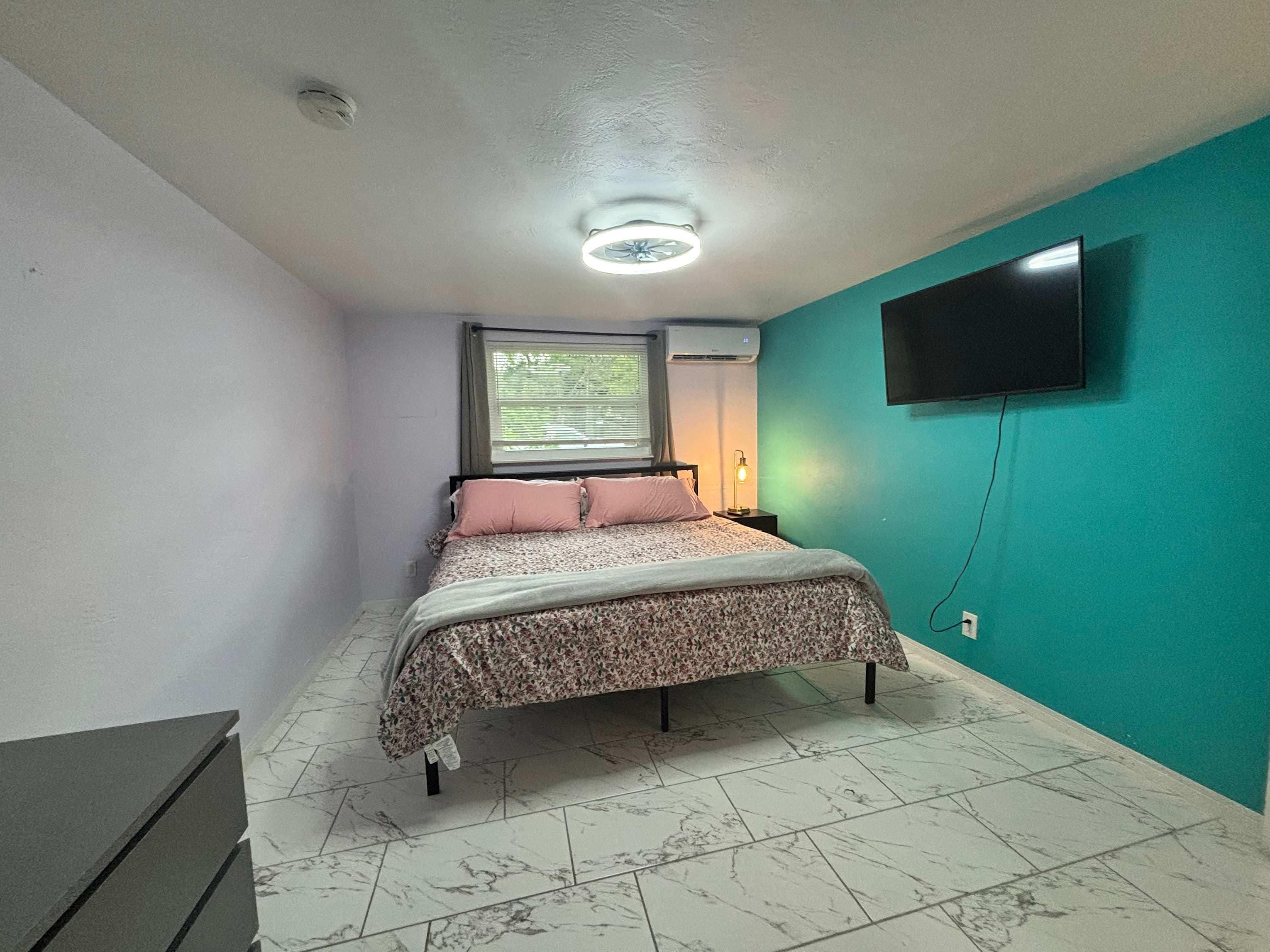 Un dormitorio con cama, televisor en la pared y ventana con persianas.