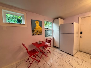Una cocina compacta con refrigerador, una mesa de comedor con sillas, un ventanal con una planta y un cuadro en la pared.