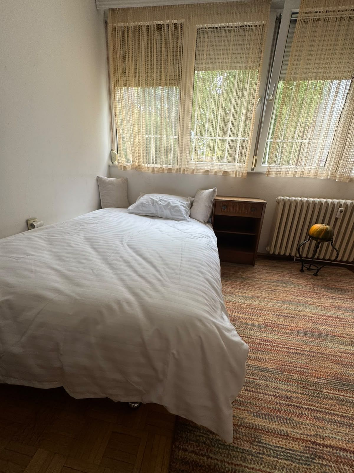 A bedroom with a bed, a nightstand, a radiator, and a window with curtains.