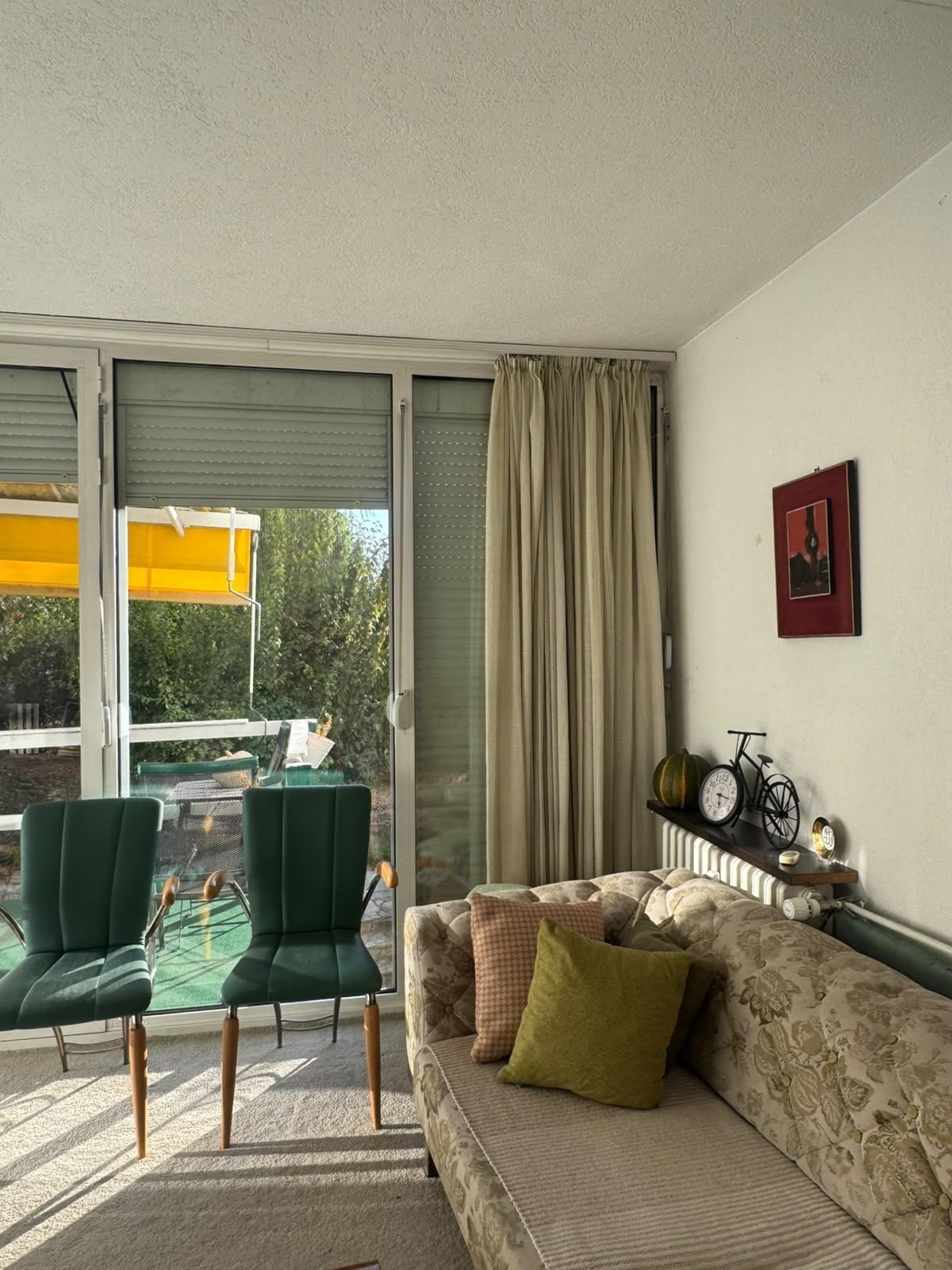 A living room with a sofa, two chairs, a radiator, a window with curtains, and a wall-mounted clock.