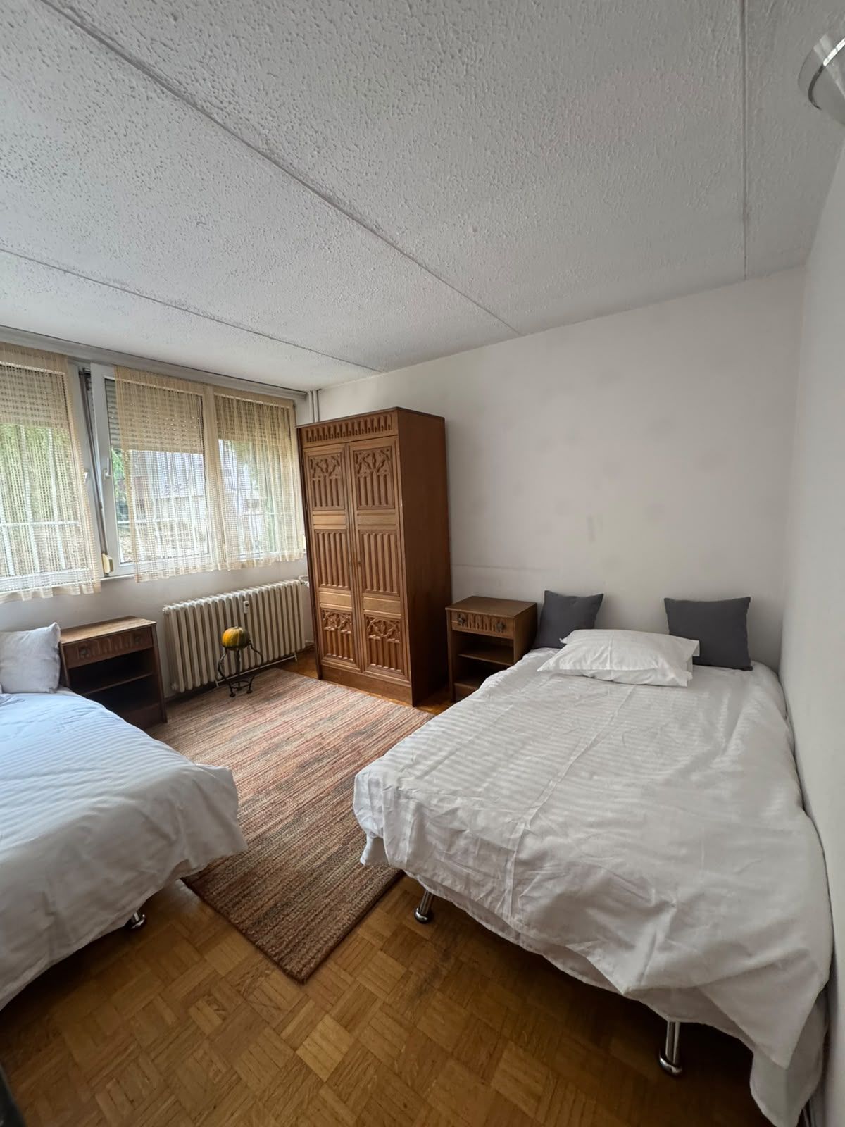 Ein Schlafzimmer mit zwei Betten, einem Holzschrank, einem Heizkörper und einem Fenster mit Vorhängen.
