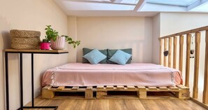Ein Schlafzimmer mit einem Bett aus Holzpaletten, einem kleinen Tisch mit Pflanzen und Blick auf den Himmel durch ein Oberlicht.