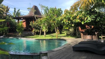 A swimming pool with a wooden deck, surrounded by lush greenery and a traditional thatched-roof structure.