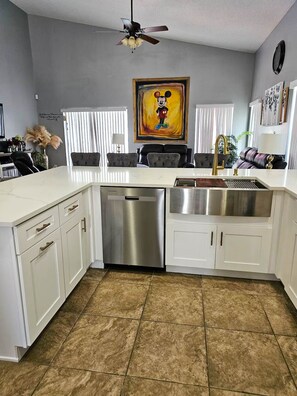 A modern kitchen with white cabinetry, a stainless steel dishwasher, and a large window with blinds. - Disney Pool Escape | Game Room + Sleeps 14 (Davenport)