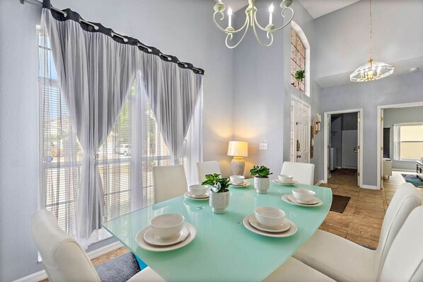 A dining room with a glass table set for four, white chairs, a chandelier, and a view through sheer curtains. - Disney Pool Escape | Game Room + Sleeps 14 (Davenport)
