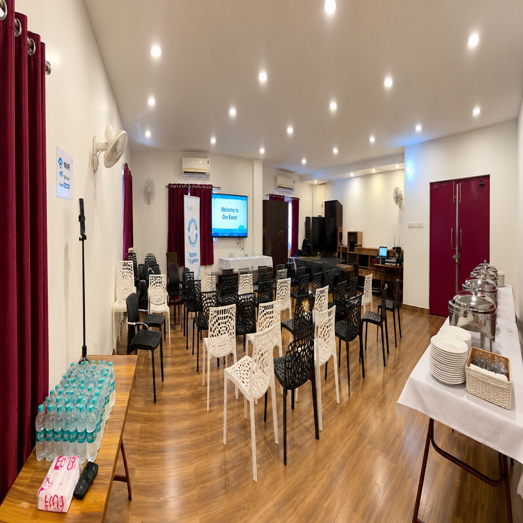 Un salón de conferencias con filas de sillas, un escenario con una pantalla y una mesa de buffet.