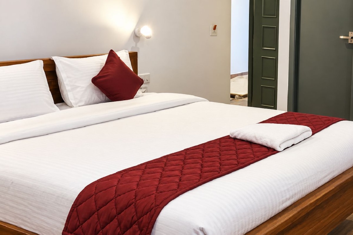 Una cama bien hecha con sábanas blancas y una colcha acolchada roja, cabecera de madera y una lámpara fijada a la pared.