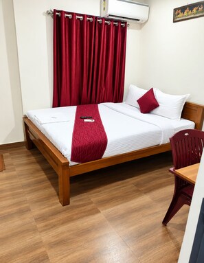 Una cama bien hecha con una manta y almohadas rojas, un cabecero de madera, una silla roja y un aire acondicionado instalado en la pared.