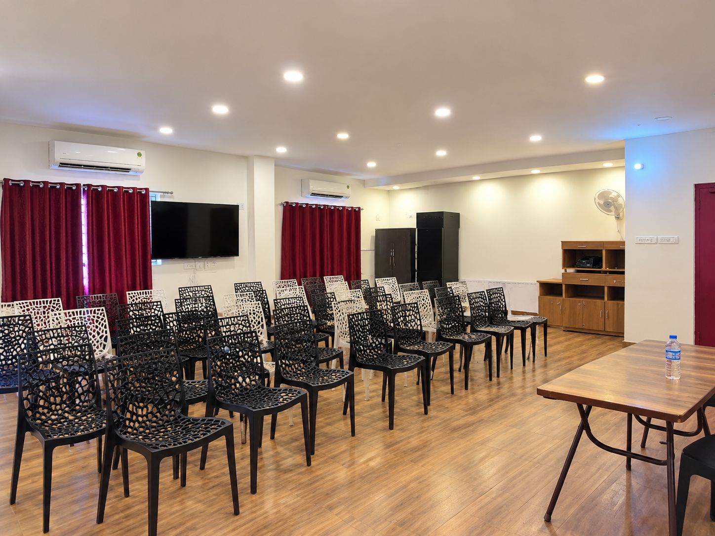 A conference room with rows of chairs, a table, an air conditioning unit, a television, and a fan.