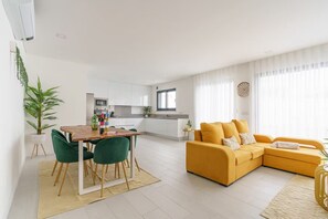 A modern living room with a yellow sofa, a dining area with a wooden table and chairs, and a kitchen in the background.