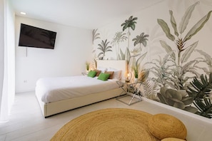 A bedroom with a bed, a wall-mounted TV, a nightstand with a lamp, and a large tropical plant mural.