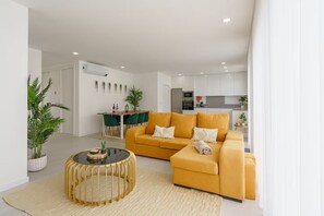 A modern living room with a yellow sofa, a round coffee table, and a dining area with green chairs.