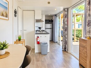 A compact kitchen with a sink, stove, and cabinets, leading to a balcony with a view of greenery. - Caravan '5-star Family' with Shared Pool, Private Terrace and Air Conditioning (Vendres)