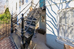 Ein schmaler, mit Backsteinen gepflasterter Gang mit einer Metalltreppe, die zu einem Gebäude mit einer blauen Tür und weißen Fensterläden führt.