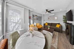 Ein modernes Wohnzimmer mit Essbereich, ausgestattet mit einem Marmortisch, Sofas mit gelben Kissen, einem Deckenventilator und einem Flachbildfernseher.
