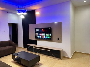 A modern living room with a flat-screen TV mounted on the wall, a dark coffee table, and a brown sofa. - Serene 2-Bed-Apt ~ 24hrs Pwr ~ Unlimited Internet (Lagos)