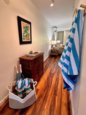 A hallway with wooden flooring, a wooden cabinet, a framed picture on the wall, a basket with bags, and a striped towel hanging on the wall. - Turtle Bay 1 Bedroom w/AC & Lanai (Kahuku)