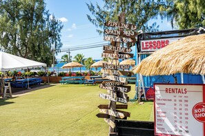 Ein hölzernes Wegweiser mit Zielortnamen und Entfernungen, ein Schild mit der Aufschrift 'LESSONS' und ein Schild mit der Aufschrift 'RENTALS', das Preise für verschiedene Aktivitäten auflistet.