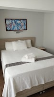 Cama bem arrumada com lençóis brancos e toalha dobrada, cabeceira de madeira, mesa de cabeceira e um quadro na parede.