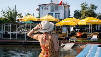 Eine Frau steht an einem Steg, in der Nähe ein Boot, im Hintergrund ein Motel und gelbe Sonnenschirme.