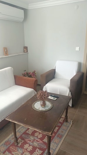 A living room with a sofa, two armchairs, a coffee table, a vase with dried flowers, a remote control, and a wall-mounted air conditioner.