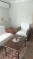 Una sala de estar con un sofá, dos sillones, una mesa de centro, un jarrón con flores secas, un control remoto y un aire acondicionado instalado en la pared.