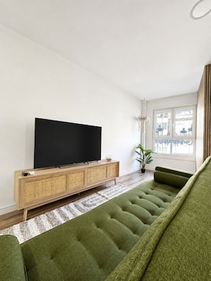 A modern living room with a flat-screen TV on a wooden console, a green sofa, and a large window with a view.
