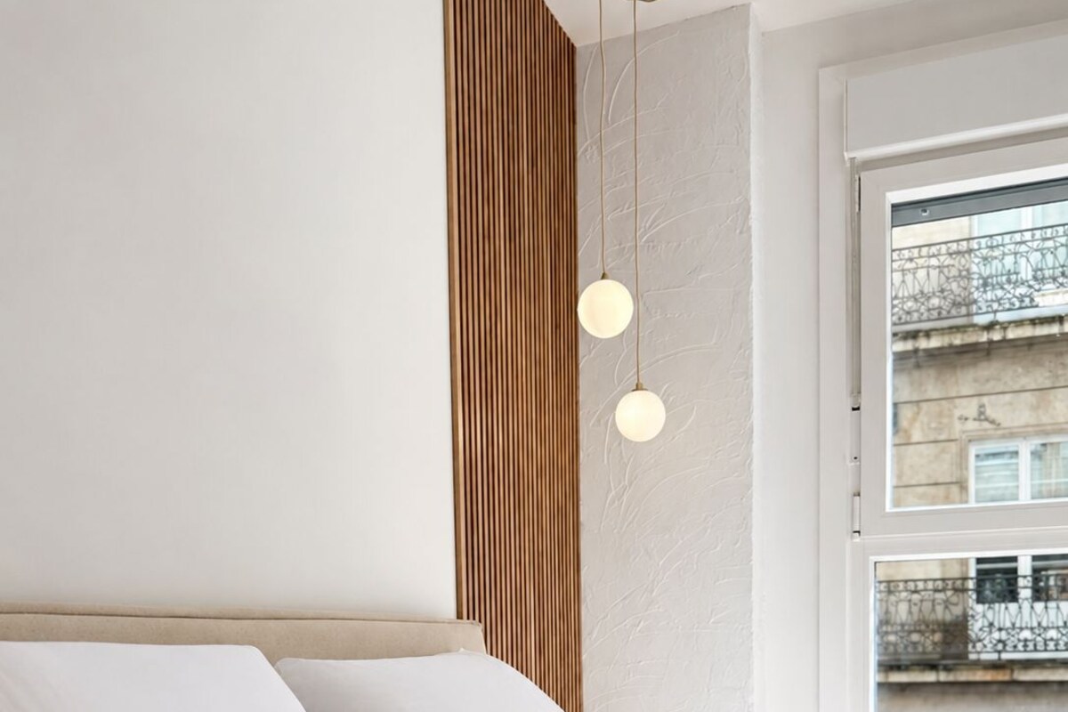 Una cama con ropa de cama blanca, un panel de pared de madera y una ventana con vista a edificios.