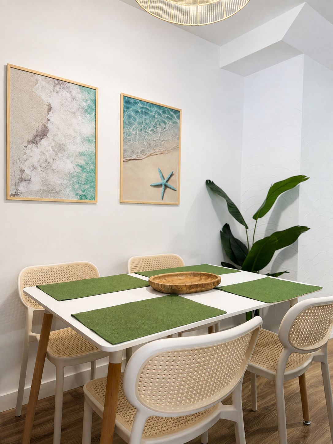 Un comedor con una mesa blanca, individuales verdes, sillas de mimbre y dos cuadros con motivos playeros.