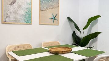 Un comedor con una mesa blanca, individuales verdes, sillas de mimbre y dos cuadros con motivos playeros.
