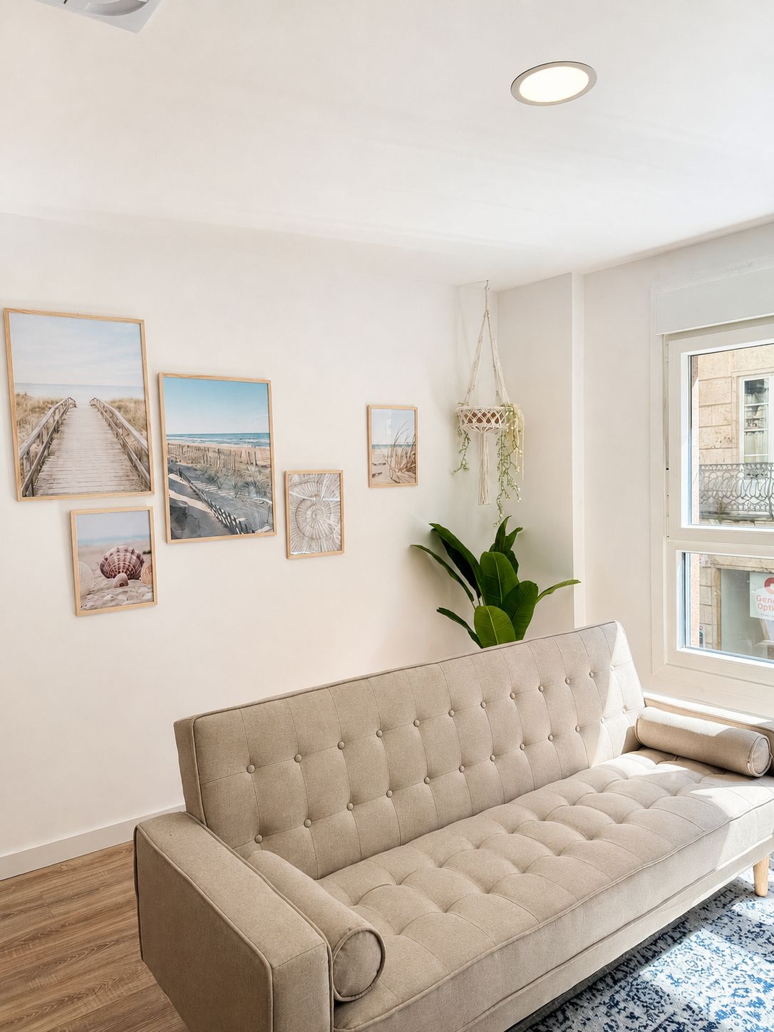 Una sala de estar con un sofá color beige, una planta de macramé colgante y cuadros con motivos playeros en la pared.