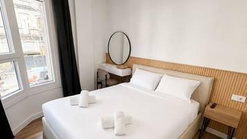 Habitación de hotel con una cama grande, una mesita de noche, un escritorio con silla, un espejo redondo y una ventana con cortinas.