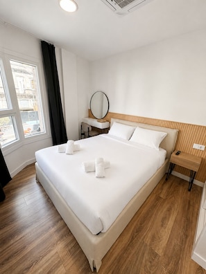 A hotel room with a large bed, a bedside table, a desk with a chair, a round mirror, and a window with curtains.