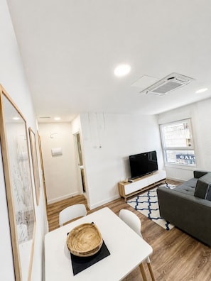 A modern living room with a flat-screen TV, a grey sofa, and a dining area with a white table and chairs.