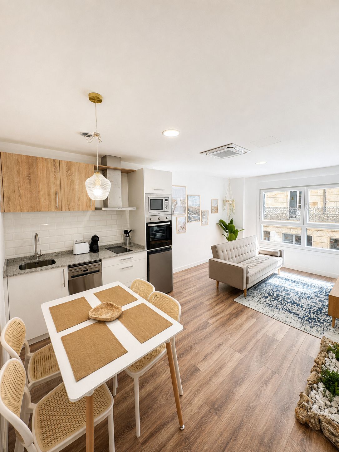 Una cocina moderna con armarios blancos, una mesa de comedor con individuales y un sofá en una sala de estar con vistas a través de amplios ventanales.