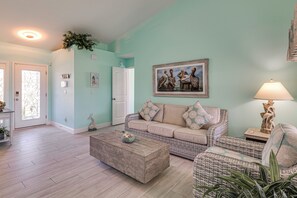 Een woonkamer met een lichtblauwe muur, een witte deur, een beige bank met patroon kussens, een rieten stoel, een houten salontafel en een lijst met een schilderij van pelikanen.