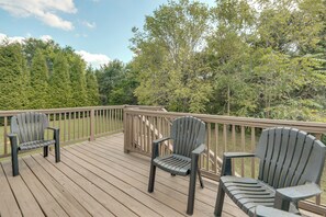 Um deck de madeira com cadeiras Adirondack e guarda-corpo com vista para uma área arborizada.