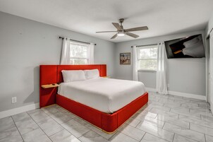 A bedroom with a red bed, white bedding, a ceiling fan, and two windows with white curtains.