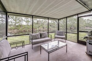 Un área de estar al aire libre con techo, que incluye un sofá, sillas y una mesa, con vistas a un paisaje natural con árboles y una masa de agua.