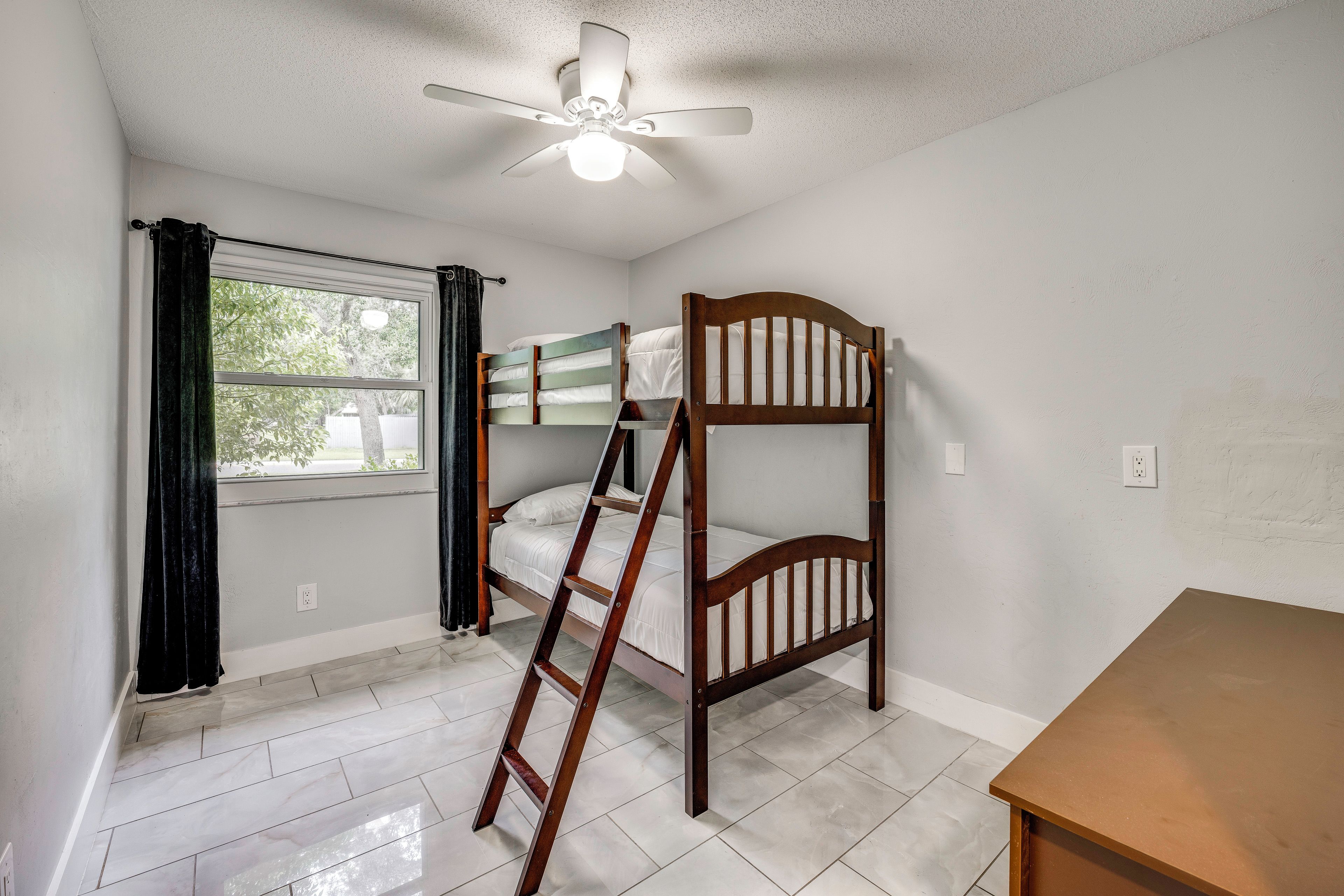 A room with bunk beds, a ceiling fan, a window with curtains, and a wooden table.