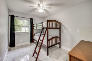 Una habitación con literas, ventilador de techo, ventana con cortinas y una mesa de madera.