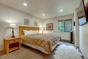 A bedroom with a wooden bed, bedside lamps, framed pictures, a window with a view of trees, and a white furry rug. - Steps to Slopes! Mammoth Mountain Condo (Mammoth Lakes)