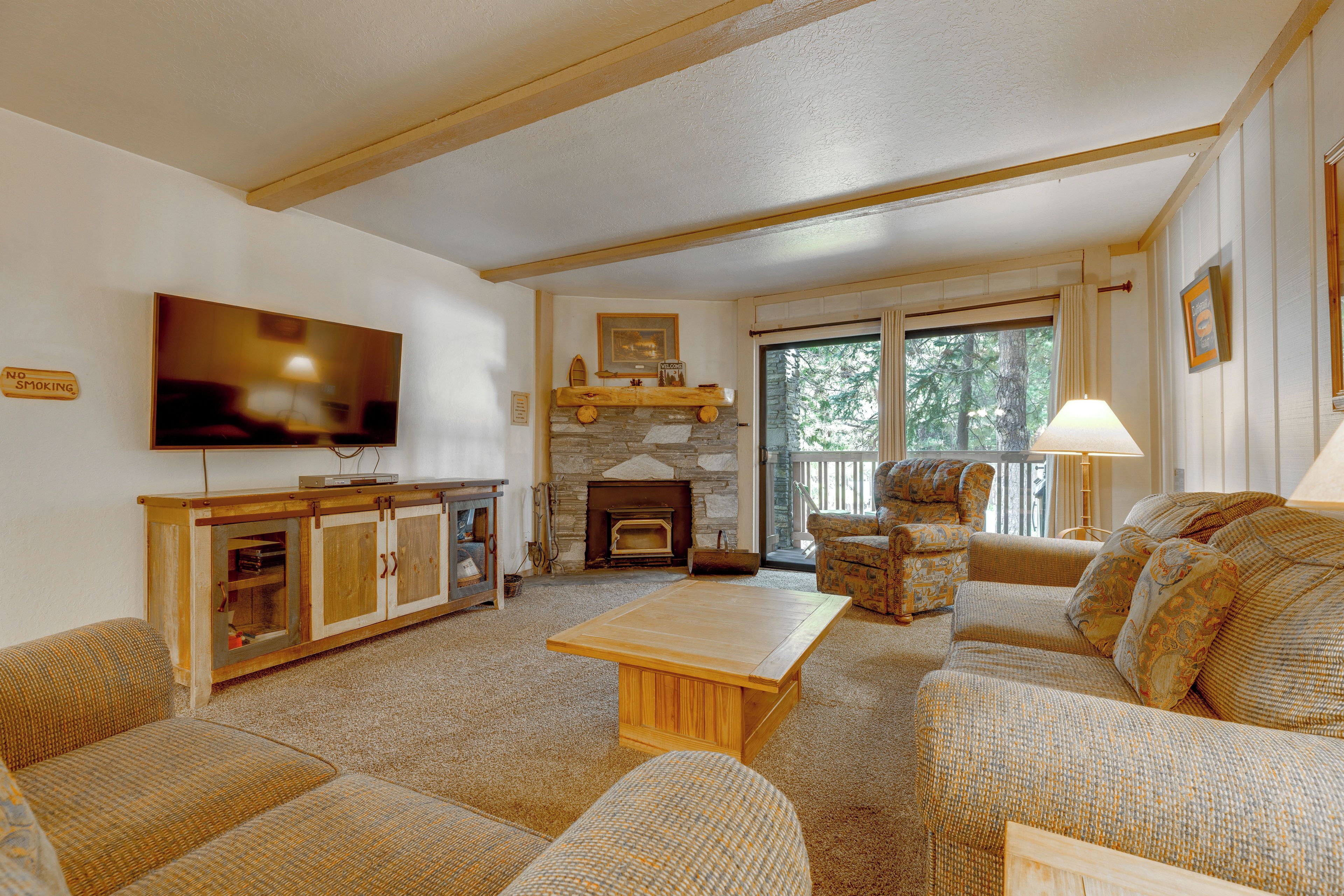 A cozy living room with a fireplace, comfortable seating, a flat-screen TV, and a large window with a view of trees.